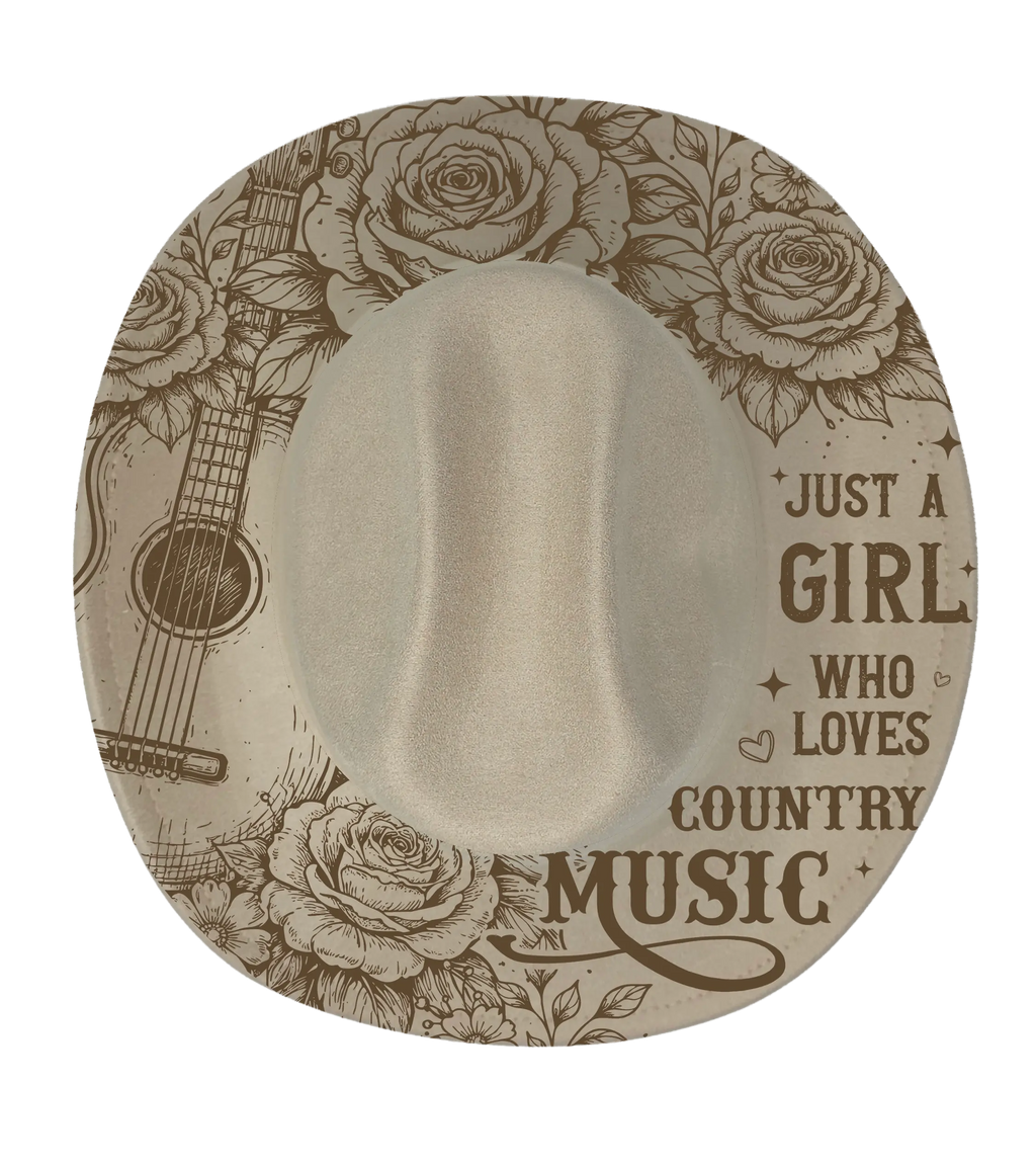 A biege cattleman hat with a guitar and roses engraved next to text that says just a girl who loves country music